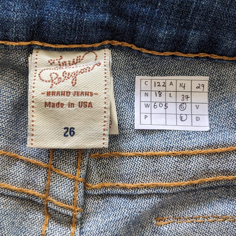 May include: A close-up of a blue denim jean label with the text "True Religion Brand Jeans Made in USA" and the size "26". A white tag with size measurements is also visible.