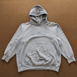 Puede incluir: Una sudadera con capucha gris con una corona negra y el texto "JEAN-MICHEL BASQUIAT" impreso en la parte delantera.