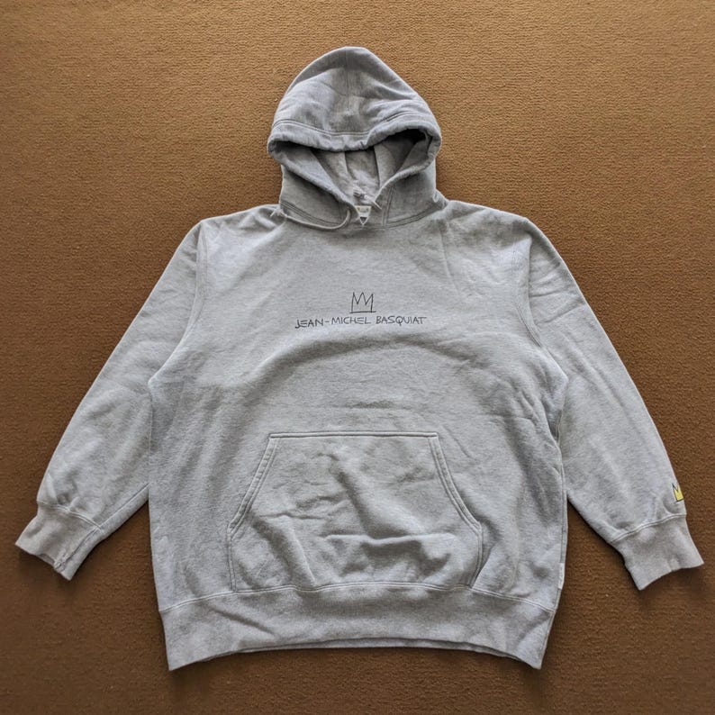 Puede incluir: Una sudadera con capucha gris con una corona negra y el texto "JEAN-MICHEL BASQUIAT" impreso en la parte delantera.
