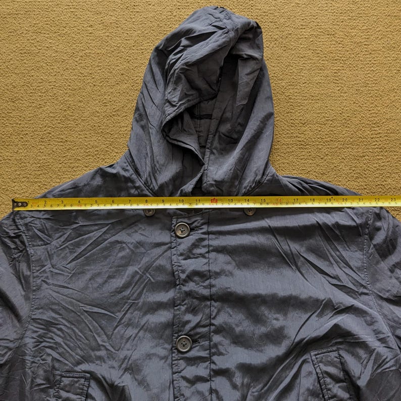 Puede incluir: Chaqueta con capucha gris oscuro con cierre de botones. La chaqueta tiene capucha y dos bolsillos. Una cinta m&eacute;trica se coloca sobre la chaqueta, mostrando su ancho. La chaqueta parece estar hecha de un material ligero.