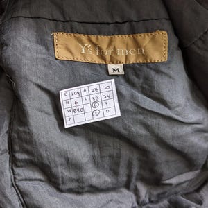 Puede incluir: Primer plano del interior de una chaqueta gris. Una etiqueta marr&oacute;n con el texto "Y's for men" est&aacute; cosida en la chaqueta. Debajo, una etiqueta blanca con n&uacute;meros y letras.