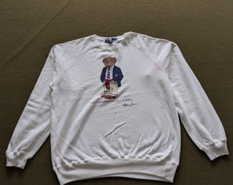May include: White long-sleeve sweatshirt featuring a graphic of a bear wearing a blazer, tie, and slacks. The text "POLO BEAR by RALPH LAUREN" is printed below the bear. The sweatshirt has a crew neck and raglan sleeves.