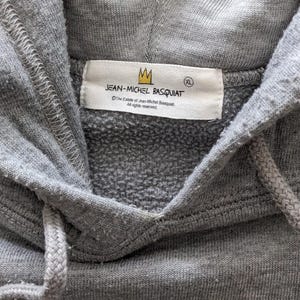 Puede incluir: Sudadera gris con capucha con una etiqueta blanca que dice "JEAN-MICHEL BASQUIAT" con un logotipo de corona y el texto "The Estate of Jean-Michel Basquiat. All rights reserved." La etiqueta tambi&eacute;n tiene una etiqueta de talla que dice "XL".