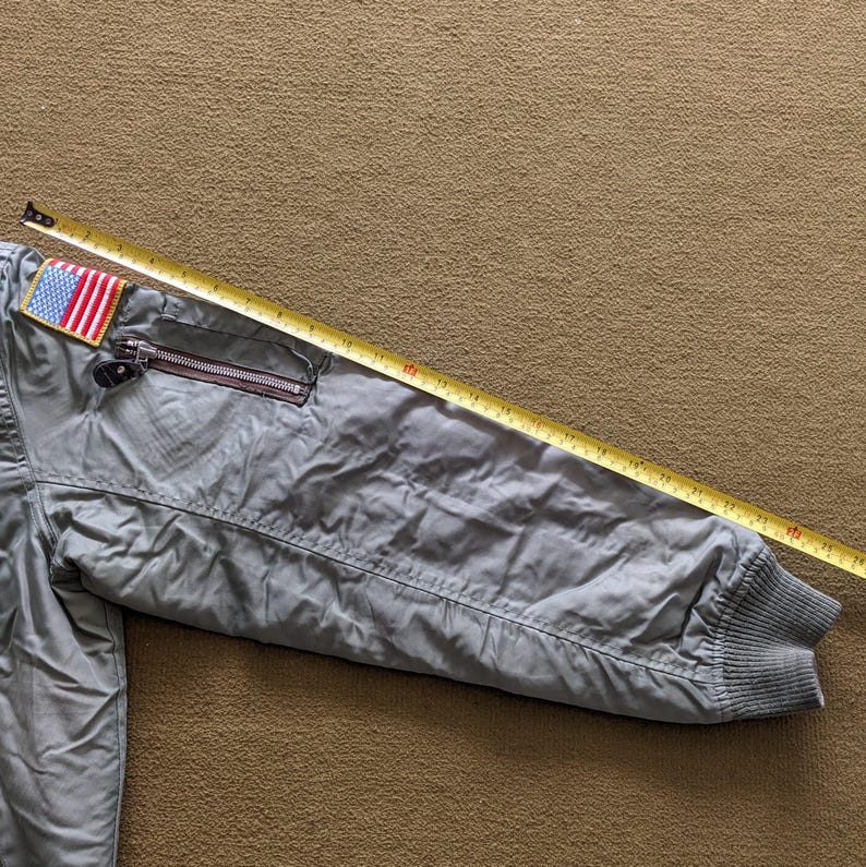 May include: A grey bomber jacket with a zipped pocket and an American flag patch on the left sleeve. A yellow measuring tape is laid across the jacket.