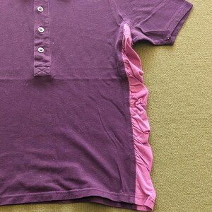 May include: A purple polo shirt with a contrasting pink ruffled panel on the side. The shirt has a classic collar, three buttons, and short sleeves. The fabric appears to be a soft, breathable material.