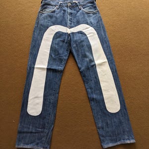 Puede incluir: Vaqueros de mezclilla azul con un diseño gráfico blanco en forma de M en cada pierna.