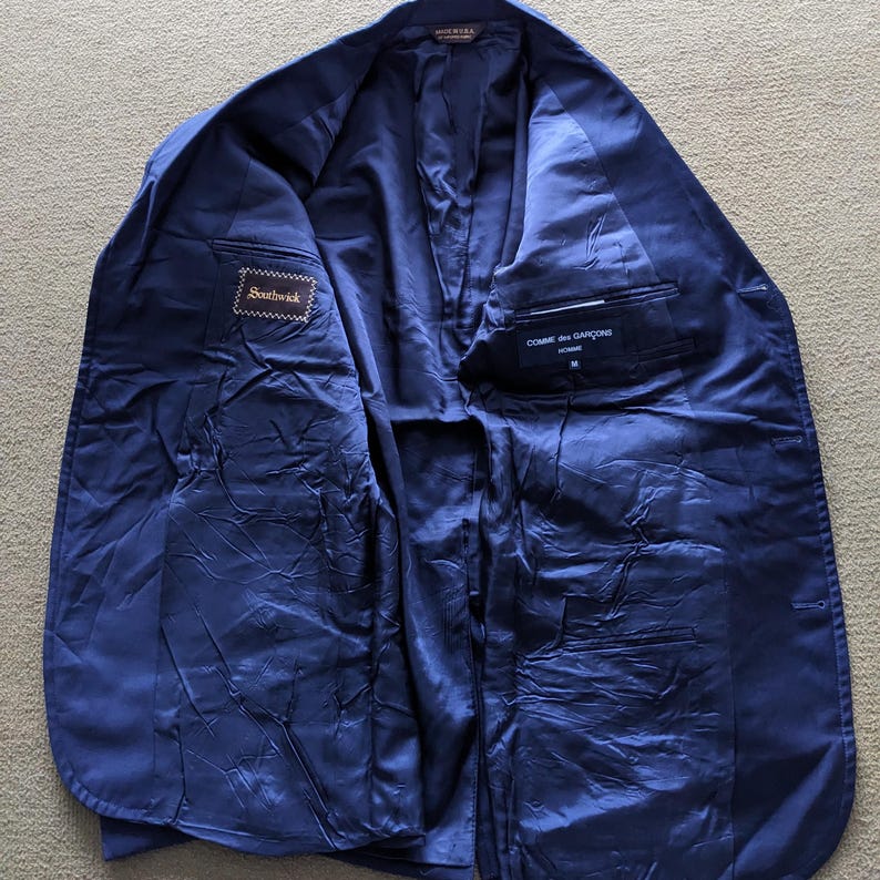 Puede incluir: Blazer azul marino abierto, que revela el forro interior y los bolsillos. El blazer tiene etiquetas que dicen "Southwick" y "Comme des Garcons". El forro es de un tono azul m&aacute;s oscuro que la tela exterior.