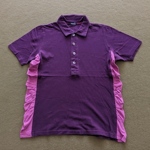 May include: A short-sleeved purple polo shirt with a contrasting pink ruffled panel on the side. The shirt has a button-up placket and a collared neckline. The fabric appears to be a soft knit.