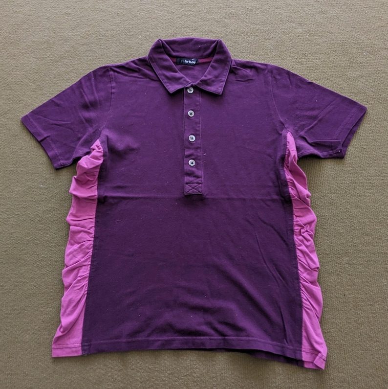 May include: A short-sleeved purple polo shirt with a contrasting pink ruffled panel on the side. The shirt has a button-up placket and a collared neckline. The fabric appears to be a soft knit.