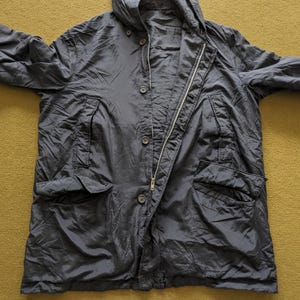 Puede incluir: Una chaqueta azul marino oscuro con capucha, cremallera y botones. La chaqueta tiene dos bolsillos delanteros y est&aacute; hecha de un material ligero. La chaqueta est&aacute; tendida sobre una superficie amarillo-verdosa.