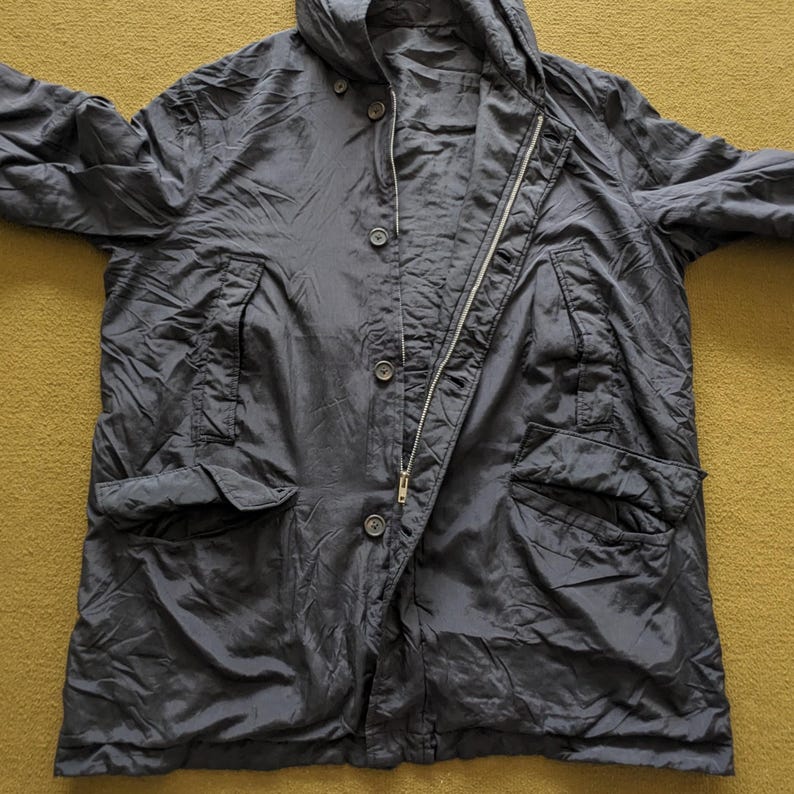 Puede incluir: Una chaqueta azul marino oscuro con capucha, cremallera y botones. La chaqueta tiene dos bolsillos delanteros y est&aacute; hecha de un material ligero. La chaqueta est&aacute; tendida sobre una superficie amarillo-verdosa.