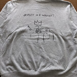 Puede incluir: Sudadera gris con un estampado gr&aacute;fico negro de una corona y tres figuras. El texto "JERSEY JOE WALCOTT" est&aacute; impreso sobre el gr&aacute;fico.