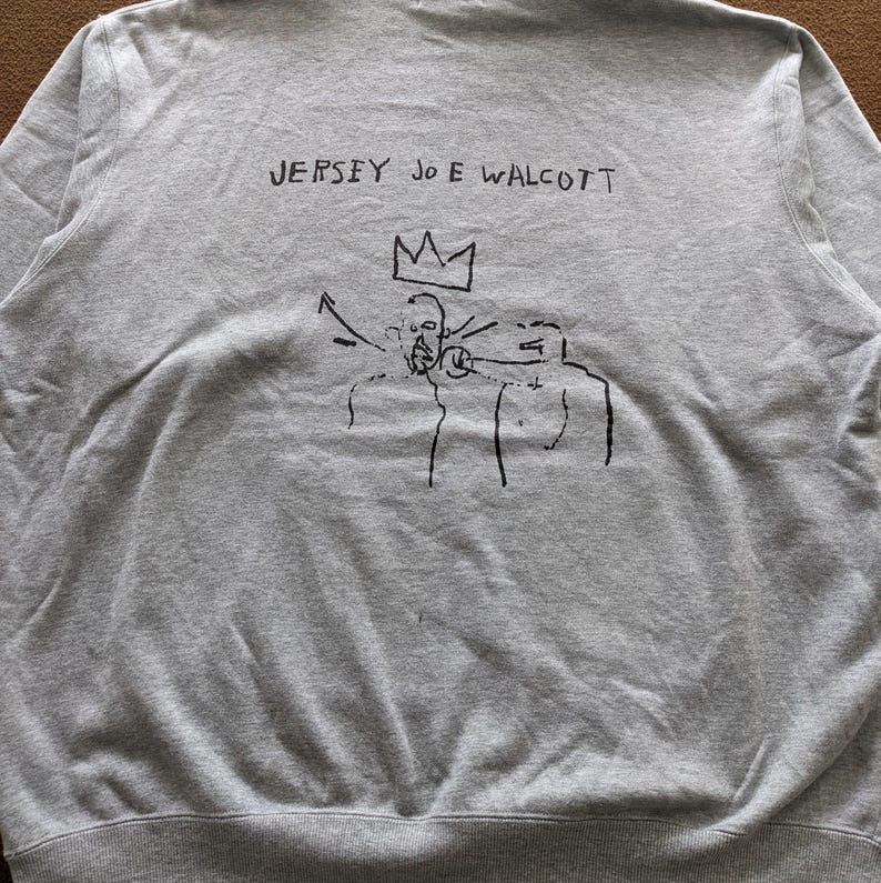 Puede incluir: Sudadera gris con un estampado gr&aacute;fico negro de una corona y tres figuras. El texto "JERSEY JOE WALCOTT" est&aacute; impreso sobre el gr&aacute;fico.