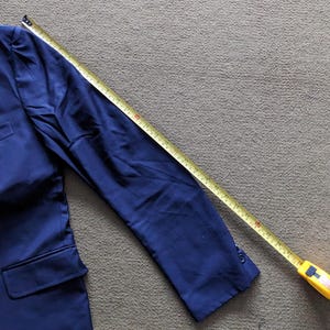 Puede incluir: Un blazer azul marino con cierre de un bot&oacute;n y un bolsillo. Una cinta m&eacute;trica amarilla se coloca sobre la manga, mostrando la longitud de la manga. El blazer es de tela lisa.