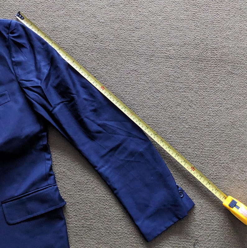 Puede incluir: Un blazer azul marino con cierre de un bot&oacute;n y un bolsillo. Una cinta m&eacute;trica amarilla se coloca sobre la manga, mostrando la longitud de la manga. El blazer es de tela lisa.