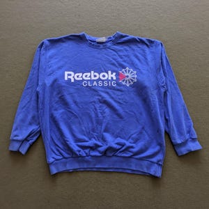 Op de afbeelding: Een blauwe Reebok Classic sweatshirt. De sweatshirt heeft het Reebok-logo in wit met een rood-blauwe afbeelding. De longsleeve crew neck shirt is gemaakt van een zacht materiaal.