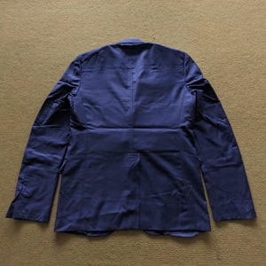 Puede incluir: Un blazer azul marino con mangas largas, visto desde la espalda. El blazer tiene un dise&ntilde;o cl&aacute;sico con solapas y pu&ntilde;os abotonados. La tela parece tener un ligero brillo, lo que sugiere un estilo formal o semi-formal.