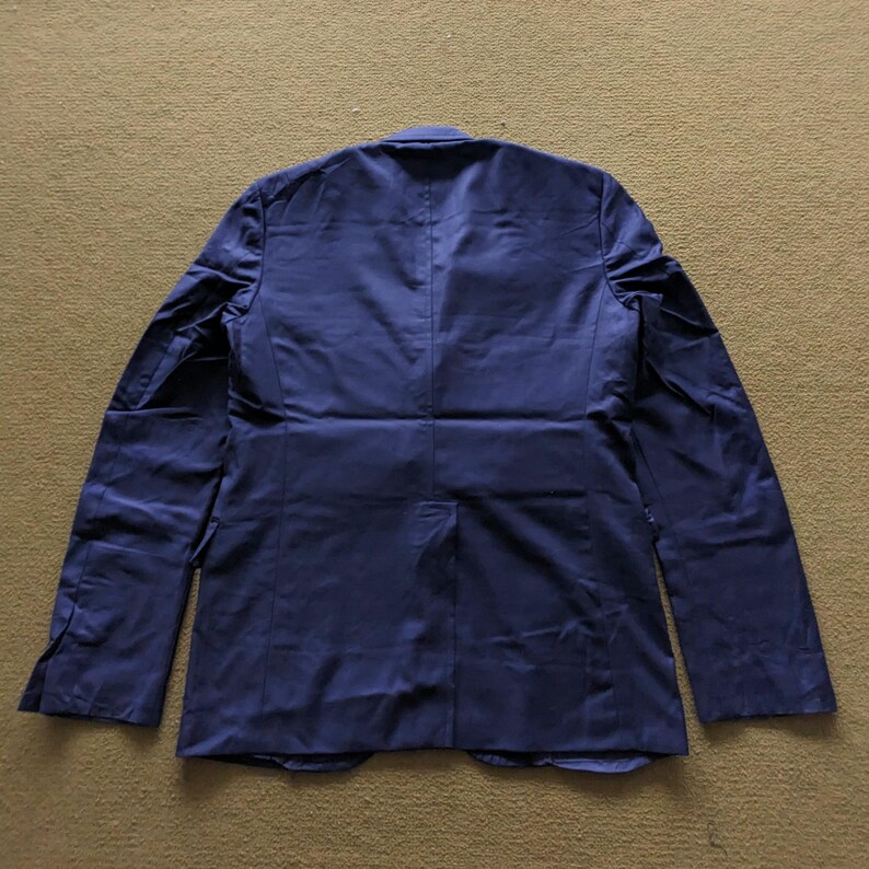 Puede incluir: Un blazer azul marino con mangas largas, visto desde la espalda. El blazer tiene un dise&ntilde;o cl&aacute;sico con solapas y pu&ntilde;os abotonados. La tela parece tener un ligero brillo, lo que sugiere un estilo formal o semi-formal.