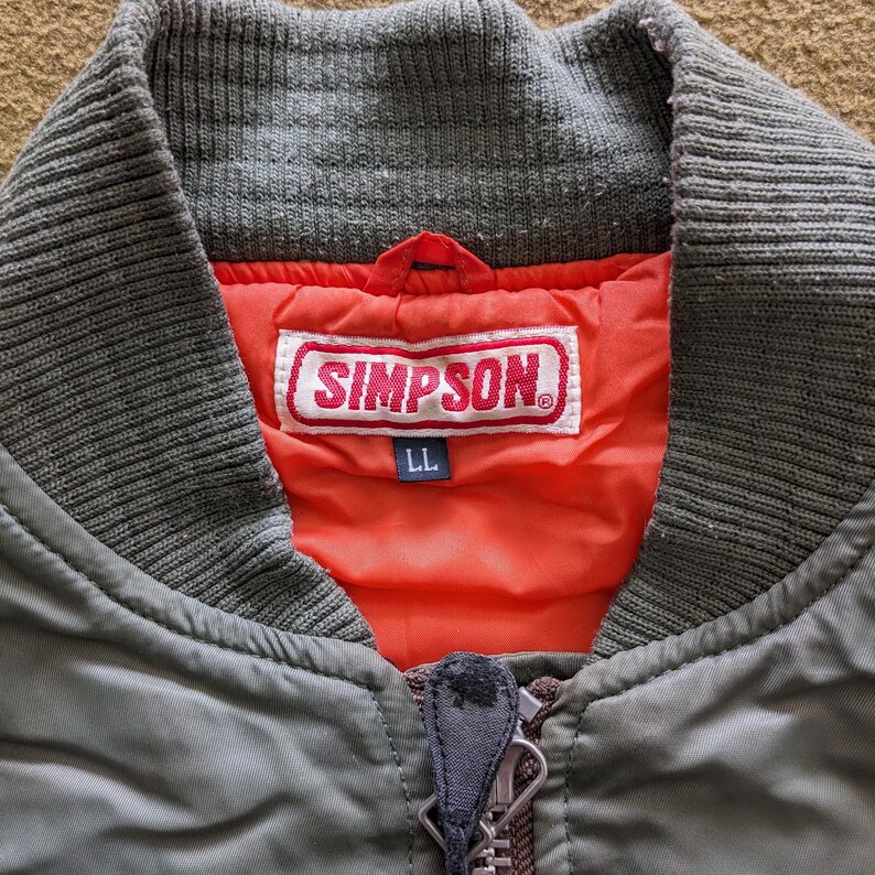 May include: A green bomber jacket with a red and white Simpson logo patch on the chest. The jacket has a zipper closure and a ribbed collar.