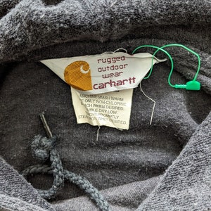 Op de afbeelding: Een close-up van een grijze Carhartt hoodie met een label waarop staat "rugged outdoor wear carhartt". Het label bevat ook wasinstructies.