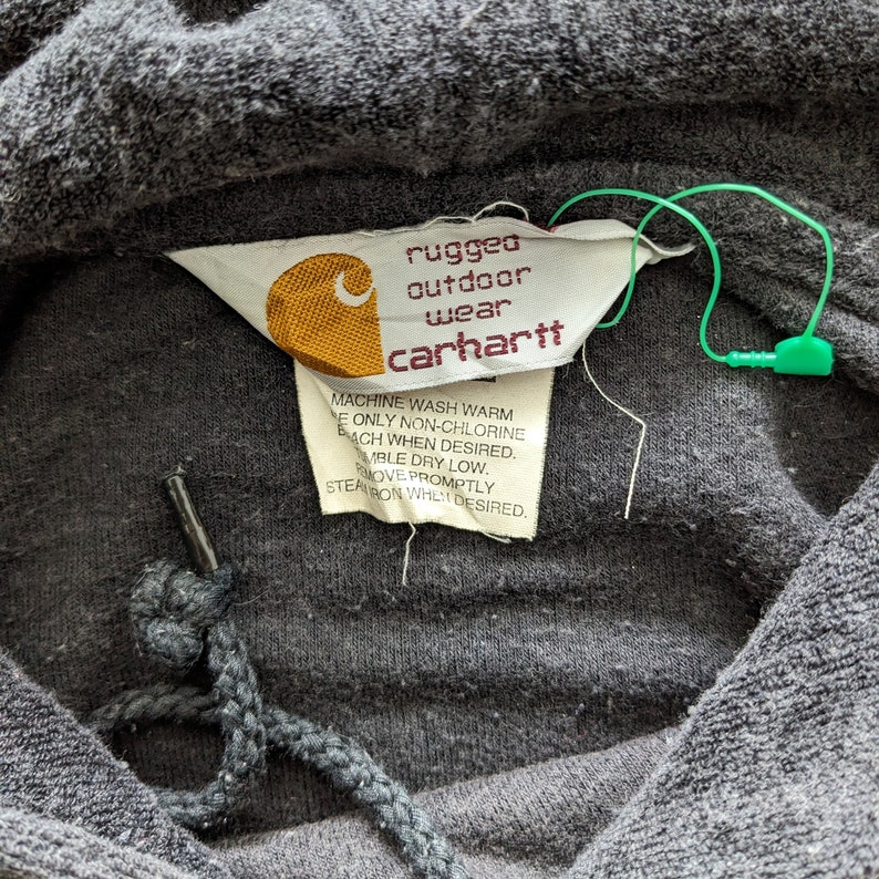 Op de afbeelding: Een close-up van een grijze Carhartt hoodie met een label waarop staat "rugged outdoor wear carhartt". Het label bevat ook wasinstructies.