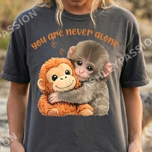 May include: Dark gray t-shirt featuring a graphic of two hugging monkeys. The orange text above the monkeys reads "you are never alone" with two small heart symbols. The shirt is a casual style.