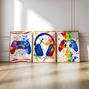 Peut inclure: Trois impressions encadrées à l'aquarelle sur le thème des jeux vidéo. L'une représente une manette rouge et bleue, une autre un casque bleu et la troisième une manette rouge, jaune et bleue. Chaque impression a des éclaboussures de peinture colorées.