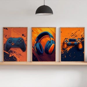 May include: Three framed art prints featuring gaming themes. The prints showcase a blue and orange color scheme with a game controller, headphones, and another controller. The artwork is displayed on a wooden shelf.