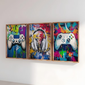 May include: A triptych of framed art prints with a gaming theme. Each print depicts a different element: a white game controller, a pair of white headphones, and another game controller. The backgrounds are vibrant with colourful graffiti-style paint splatters.