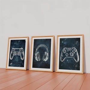 May include: Three framed prints with gaming illustrations. The prints show a game controller, headphones, and another game controller, all in white against a dark blue, starry backdrop. The frames are made of light-coloured wood.