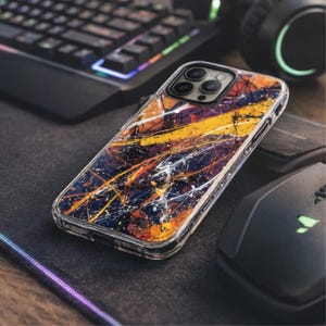Puede incluir: Funda transparente para teléfono con un diseño abstracto de salpicaduras de pintura amarilla, naranja y blanca sobre un fondo azul oscuro y morado. La funda está sobre una superficie negra cerca de un teclado y un ratón.