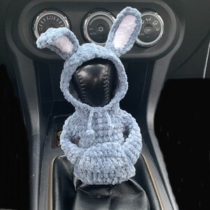 May include: A blue crocheted bunny rabbit wearing a grey hoodie is attached to a car's gear shift lever.