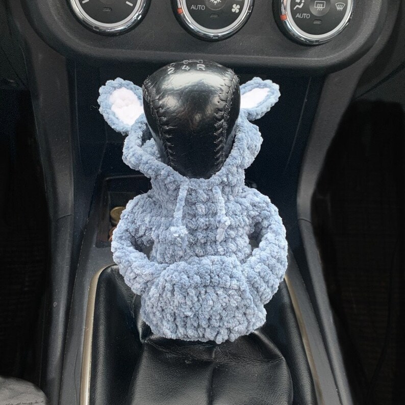 May include: A light blue crocheted car gear shift cover with white ears. The cover is shaped like a hooded sweatshirt.