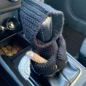 May include: A black crocheted Grim Reaper figure with a white crocheted scythe, sitting on a car's gear shift lever.