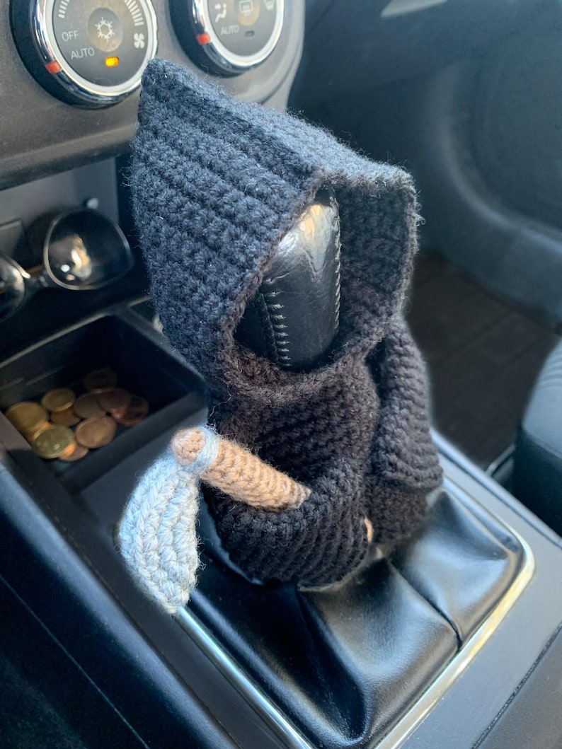 May include: A black crocheted Grim Reaper figure with a white crocheted scythe, sitting on a car's gear shift lever.