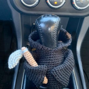 May include: A black crocheted hooded figure with a small crocheted axe in its hand. The figure is sitting on the gear shift of a car.