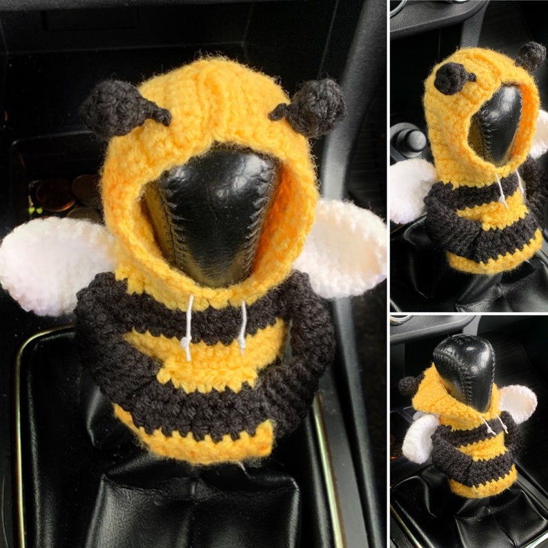 Puede incluir: Una funda de palanca de cambios de coche de ganchillo con forma de abeja con un cuerpo a rayas amarillo y negro, alas blancas y una sudadera con capucha negra.