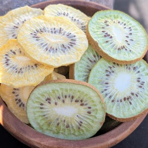 May include: A wooden bowl filled with a mix of dried kiwi slices. The fruit pieces are circular with visible dark seeds and a white centre, featuring a colour palette of pale yellow and light green with brown fuzzy edges on the green slices.