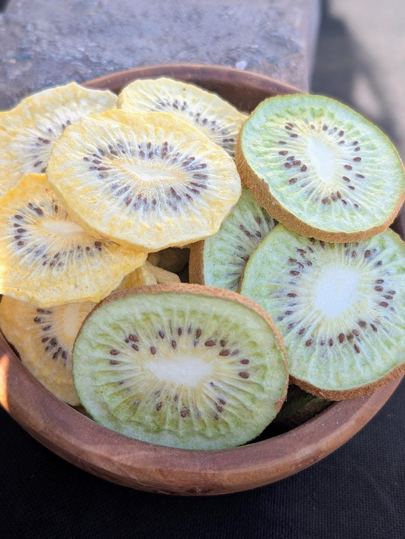 May include: A wooden bowl filled with a mix of dried kiwi slices. The fruit pieces are circular with visible dark seeds and a white centre, featuring a colour palette of pale yellow and light green with brown fuzzy edges on the green slices.