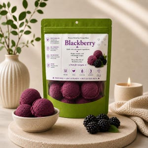 Może przedstawiać: Zielona torebka stojąca z liofilizowanymi kawałkami lodów jeżynowych. Etykieta zawiera tekst "Blackberry", "Freeze Dried Ice Cream Bites" oraz "Made with only organic ingredients". Kilka fioletowych, okrągłych kawałków lodów jest widocznych przez przezroczyste okienko oraz w małej miseczce, obok świeżych jeżyn.