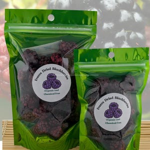 May include: Two green, resealable bags filled with freeze-dried blackberries. Each bag has a white label with a purple blackberry graphic and the text "Organic and Chemical Free". The background shows a blurred image of fresh blackberries.