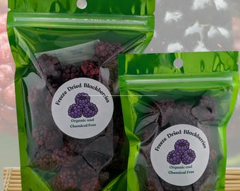 May include: Two green, resealable bags filled with freeze-dried blackberries. Each bag has a white label with a purple blackberry graphic and the text "Organic and Chemical Free". The background shows a blurred image of fresh blackberries.