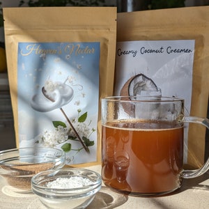 Pu&ograve; includere: Due sacchetti di carta marrone con etichette di prodotto. Il sacchetto a sinistra ha un'etichetta bianca con il testo "Heaven's Nectar" e una grafica di un fiore bianco con un vortice bianco. Il sacchetto a destra ha un'etichetta bianca con il testo "Creamy Coconut Creamer" e una grafica di una noce di cocco. Una tazza di vetro piena di un liquido marrone si trova davanti ai sacchetti. Due piccole ciotole di vetro si trovano davanti alla tazza, una piena di polvere marrone e l'altra di polvere bianca.