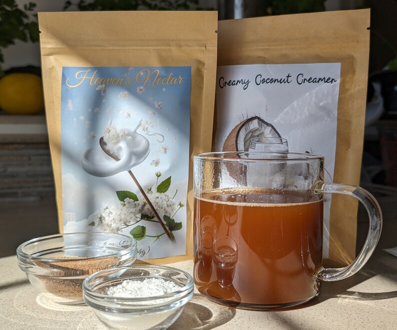 Pu&ograve; includere: Due sacchetti di carta marrone con etichette di prodotto. Il sacchetto a sinistra ha un'etichetta bianca con il testo "Heaven's Nectar" e una grafica di un fiore bianco con un vortice bianco. Il sacchetto a destra ha un'etichetta bianca con il testo "Creamy Coconut Creamer" e una grafica di una noce di cocco. Una tazza di vetro piena di un liquido marrone si trova davanti ai sacchetti. Due piccole ciotole di vetro si trovano davanti alla tazza, una piena di polvere marrone e l'altra di polvere bianca.