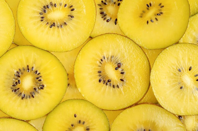 May include: A collection of sliced golden kiwi fruit arranged in a dense pattern. Each circular slice displays a vibrant yellow flesh with a central white core and a ring of small, dark black seeds embedded within the juicy, textured fruit pulp.