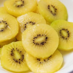 May include: A white bowl filled with multiple slices of golden kiwi fruit. Each circular slice features a bright yellow, translucent flesh with a central ring of small, dark, edible seeds arranged in a radial pattern throughout the fruit.
