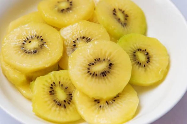 May include: A white bowl filled with multiple slices of golden kiwi fruit. Each circular slice features a bright yellow, translucent flesh with a central ring of small, dark, edible seeds arranged in a radial pattern throughout the fruit.