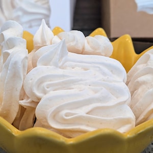 Może przedstawiać: Zbliżenie białych bezów w żółtej misce w kształcie płatka. Beza ma wzór w kształcie wiru i lekką, przewiewną konsystencję. Misa jest jasnożółta, kontrastująca z białymi ciasteczkami. W tle pudełko z podobnymi ciasteczkami.