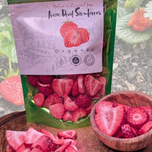 May include: A packet of freeze-dried strawberries with the text "Nature in its purest form". The packet is green and white, with a drawing of strawberries. Sliced strawberries are in a wooden bowl and scattered on a wooden surface. The product is labelled organic, gluten-free, and non-GMO.