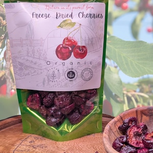 May include: A green resealable bag filled with freeze-dried cherries. The bag has a label that says "Nature in its purest form Freeze Dried Cherries Organic Gluten Free Non GMO." The bag is sitting on a wooden surface with a bowl of freeze-dried cherries next to it.